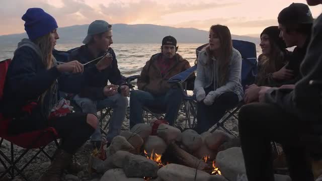 Group of millenials gathered around campfire at sunset