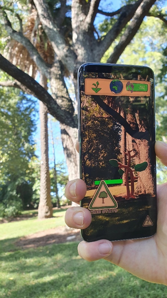 A hand holds a smartphone up against tall sunlit trees in a park; the phone screen shows the Virtual Footprints app, where a cartoon-style digital tree is overlaid onto the real trees behind it. There are on-screen icons: at the top, from left to right, are a tree planting icon, a globe indicating the player's general location, and a carbon footprint counter on green reading "-2592"; on the bottom left a button labelled "TREE STATS"; and next to the cartoon tree a full green health bar.