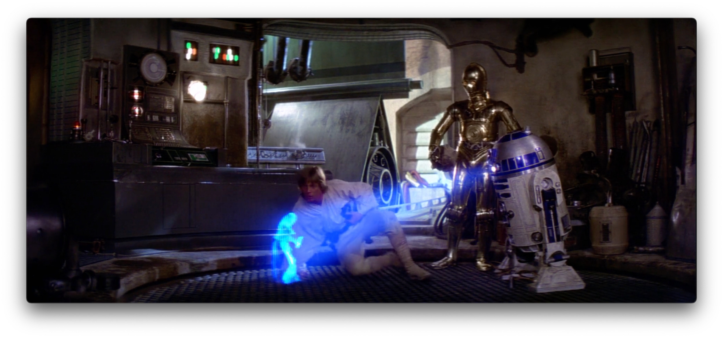 Alt text: In a still taken from A New Hope, the aluminium, white, and blue droid Artoo-Detoo uses a bright white projector positioned on his domed head to play a blue holographic video of Princess Leia, whose indistinct blue-toned figure occupies a central place in the shot. Behind the hologram, Luke Skywalker watches from a seated position on the floor, with the gold humanoid droid See-Threepio standing nearby.