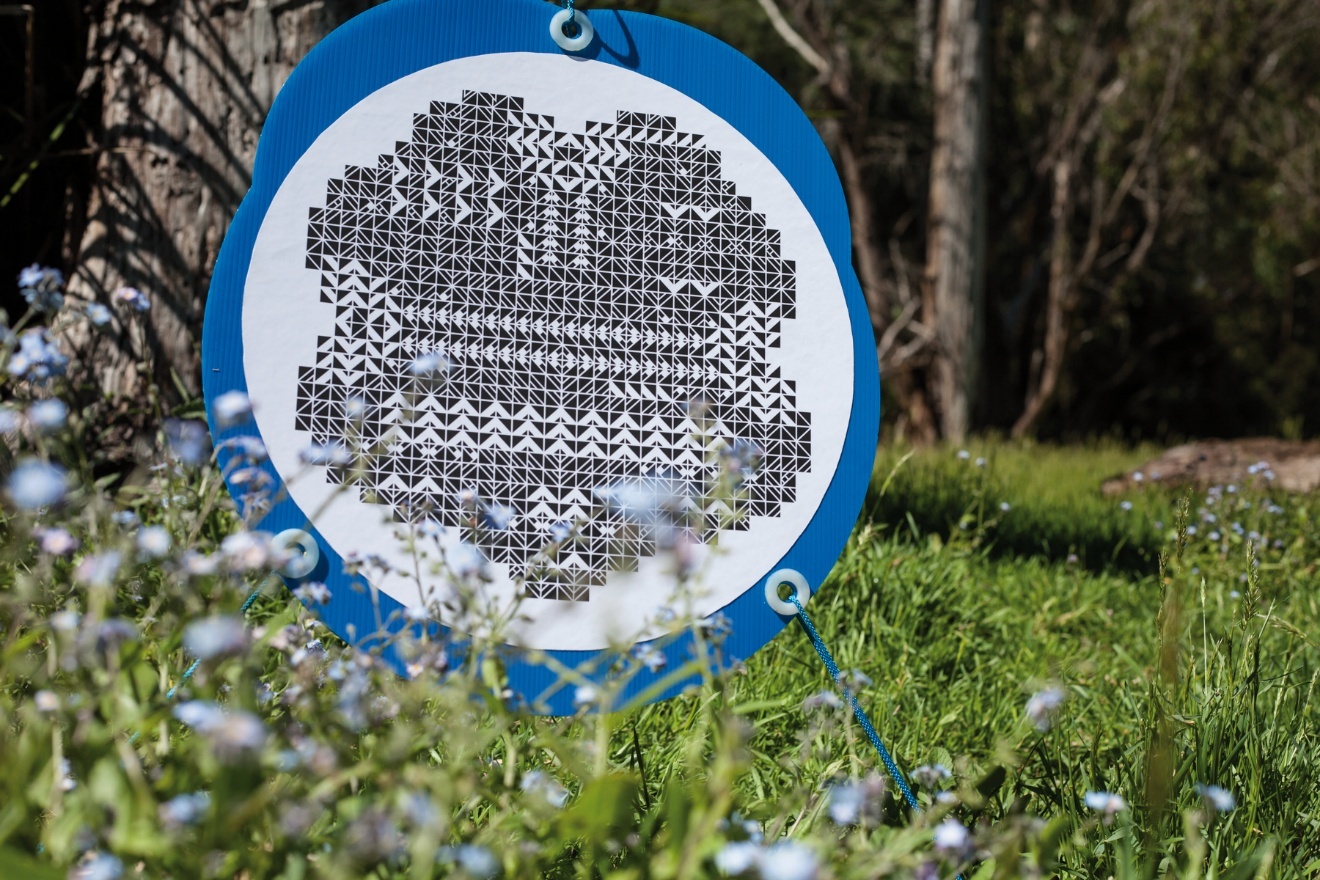 A circular marker with a blue border and a white centre, printed with a dense black-and-white pattern of small triangles forming an abstract design. It is mounted at ground level on a stake among grass and pale wildflowers, with bushland behind.