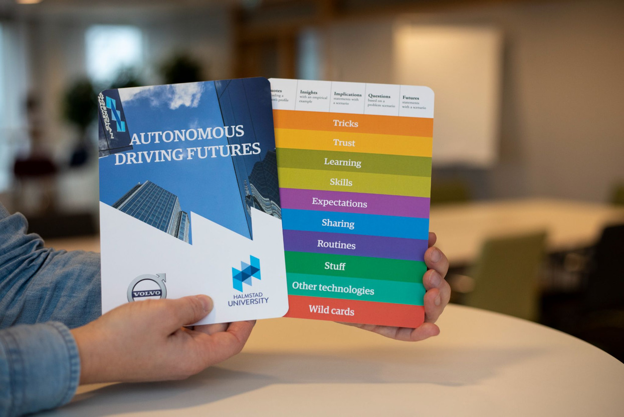Hands holding two cards, one says "Autonomous Driving Futures" and the other has many-colored stripes witht eh words: tricks, trust, learning, skills, expectations, sharing, routines, staff, other technologies, wild cards