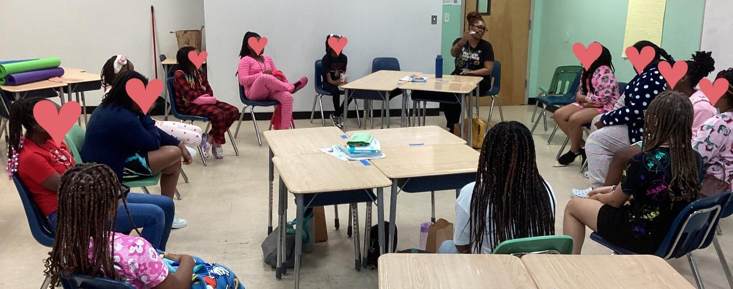 Photograph of a classroom setting with students seated in a circle around desks, engaging in a Sista Circle. Students' faces are covered with heart-shaped icons for privacy.
