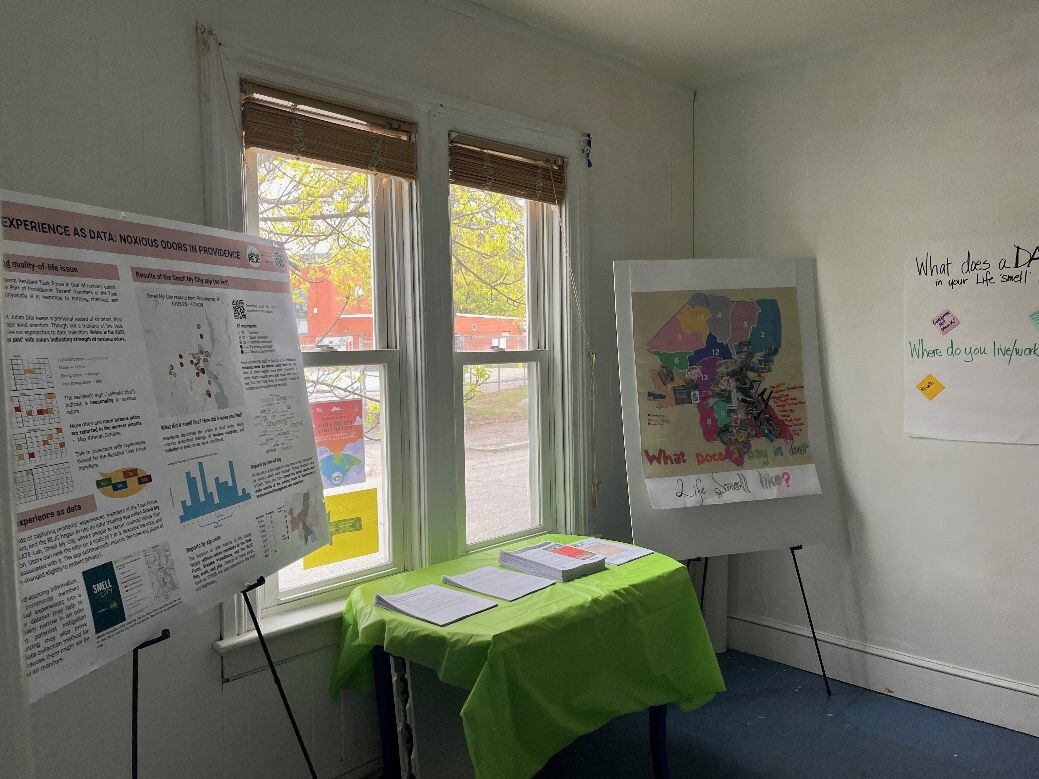 Two display stands, one with a research poster, the other of the Smellage with overlays, on either side of a table with project literature