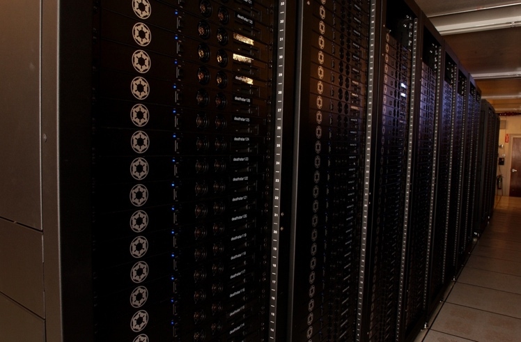  Alt text: Rows of metal cabinets recede from the foreground into the distance in a warehouse-like space. Each cabinet comprises floor-to-ceiling stacks of black computer hardware, with each unit in the stack featuring a printed logo, blue LEDs, and a variety of switches and dials.