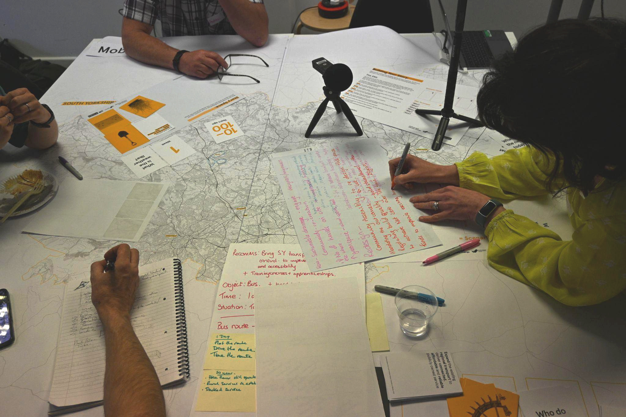 Photograph showing a table with writing materials, scenario-making cards, a map, recording equipment, and people's hands writing around the table.