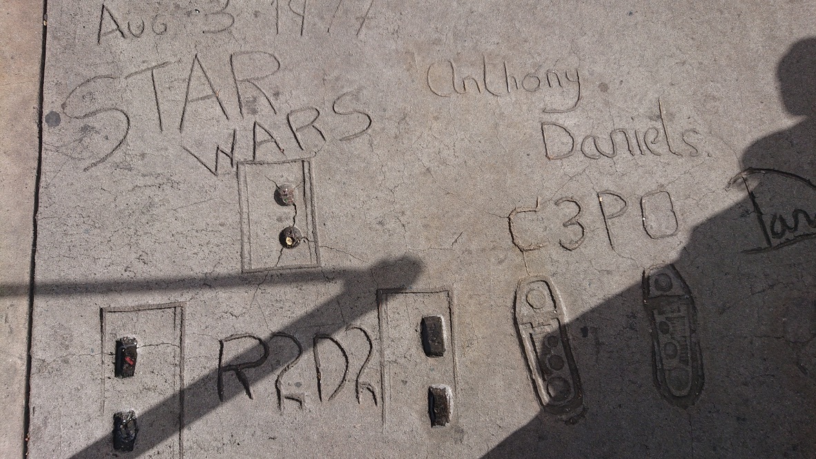 Alt text: A bird's eye view of a grey cement sidewalk that has two pairs of robot-shaped feet imprinted into it. There is text etched beside each set of set footprints: on the left is 'R2D2', and on the right 'Anthony Daniels C3PO'. Above the prints more text reads 'Aug 3 1974 Star Wars.'