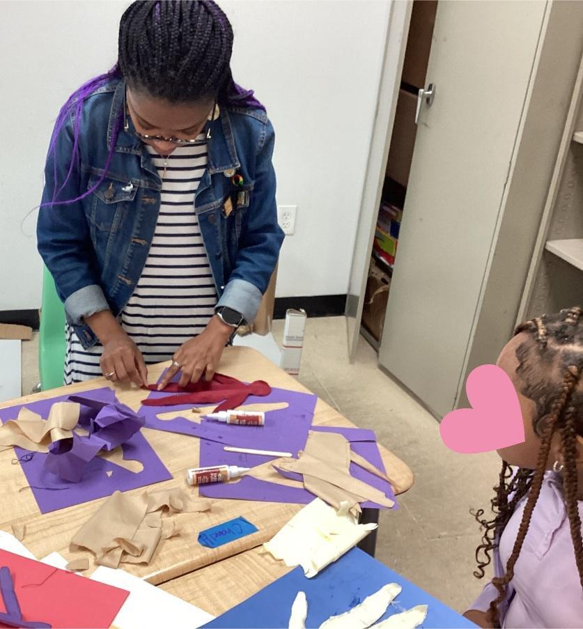 Photograph of a Black women and a Black girl engaged in creating a soft barbie doll at a table with purple and blue construction paper, and glue bottles.