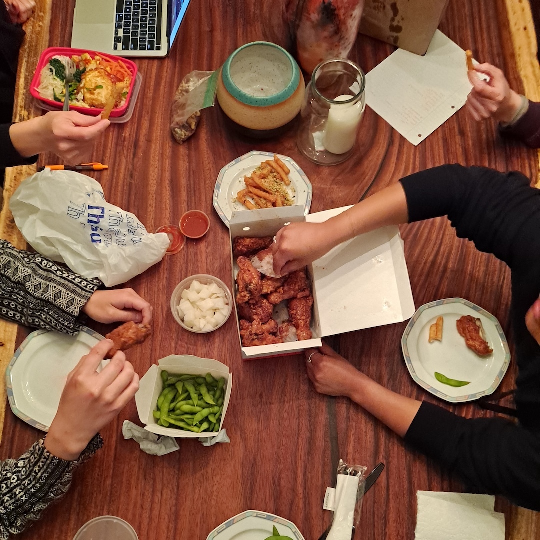 A photograph of a dining table, dishes of food in the middle surrounded by papers, a laptop, and many hands eating, sharing, and working.