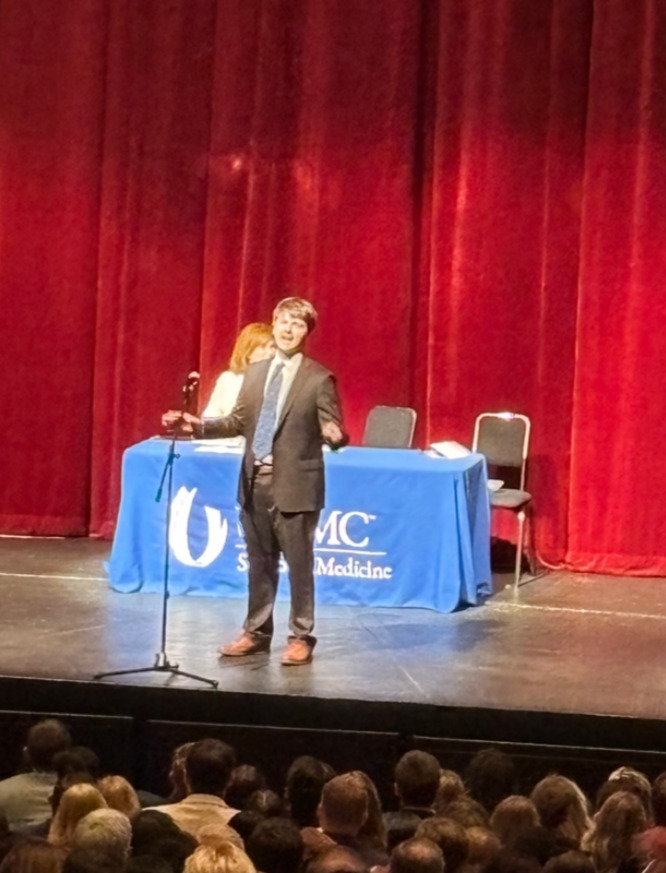 A person in suit standing on stage with a microphone Description automatically generated