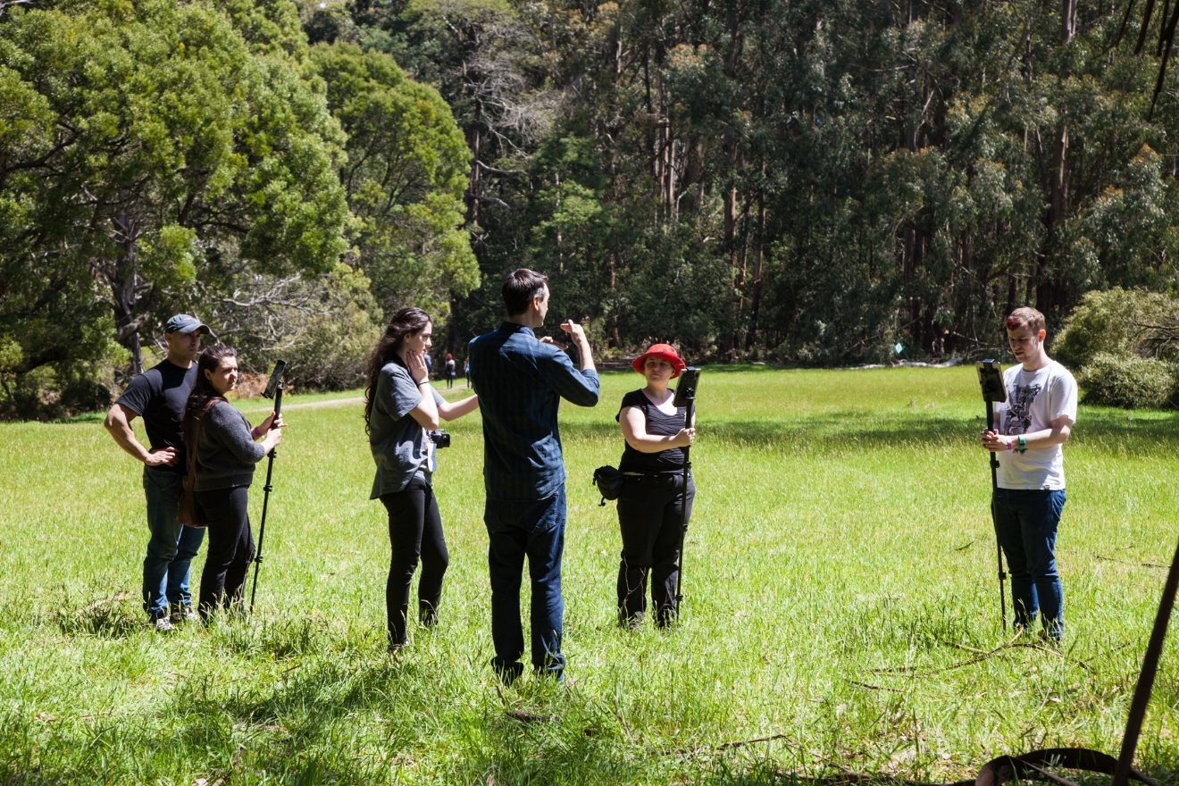 Six people stand in a loose group in a sunlit grassy clearing, bushland behind them. Several hold tablets mounted on tall monopods; one in the centre holds up a tablet as if showing it to the others. They appear to be taking part in the Epiphyte bushwalk.
