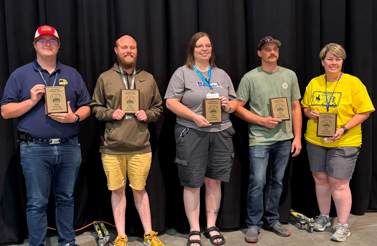 Five individuals stand in front of a black curtain, each holding a plaque for the 2026 Wyoming School Bus Rodeo.