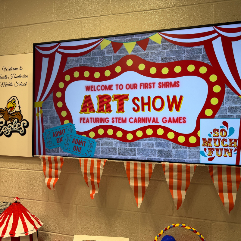 A digital screen displays 'Welcome to Our First SHRMS Art Show Featuring STEM Carnival Games' with circus-themed graphics.