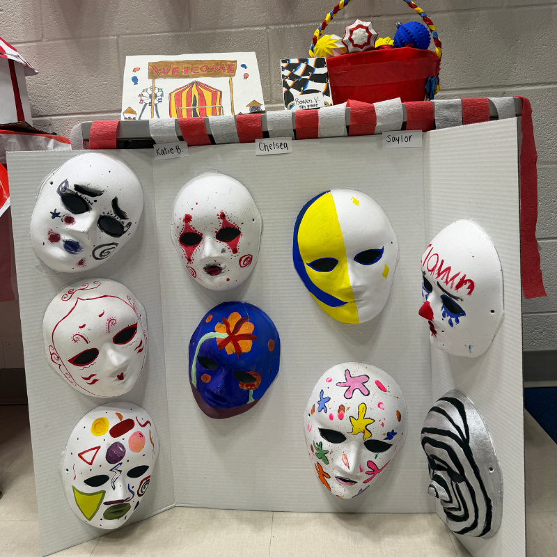 A display of colorful, hand-painted masks, some with clown-like designs, are mounted on a white board.