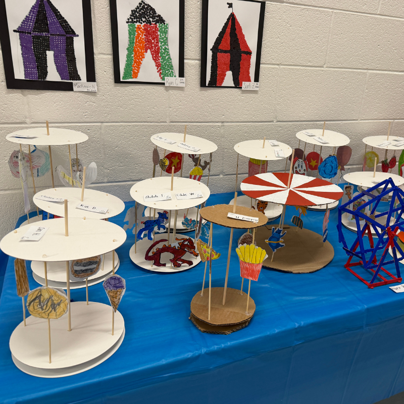 A display of student-made carousel models with colorful circus-themed artwork above.
