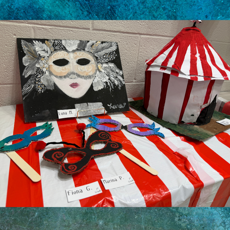 A display of student artwork featuring a painted masquerade mask, a circus tent model, and colorful handmade masks on a red and white striped tablecloth.