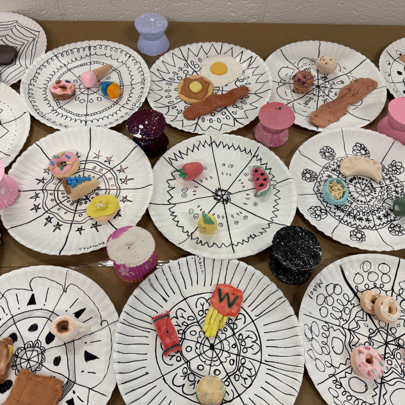 A collection of paper plates decorated with black mandala patterns, each adorned with various small clay food items.
