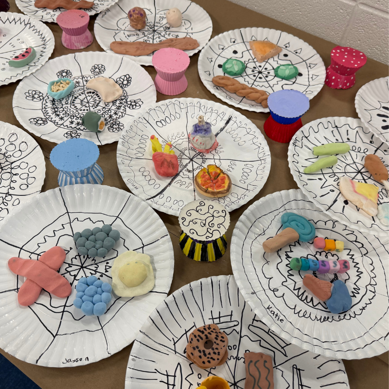 A collection of paper plates decorated with black ink designs, each holding colorful clay creations.