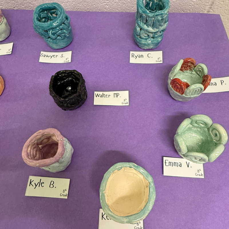 A display of colorful, handmade ceramic pots created by 8th-grade students, each labeled with a name.