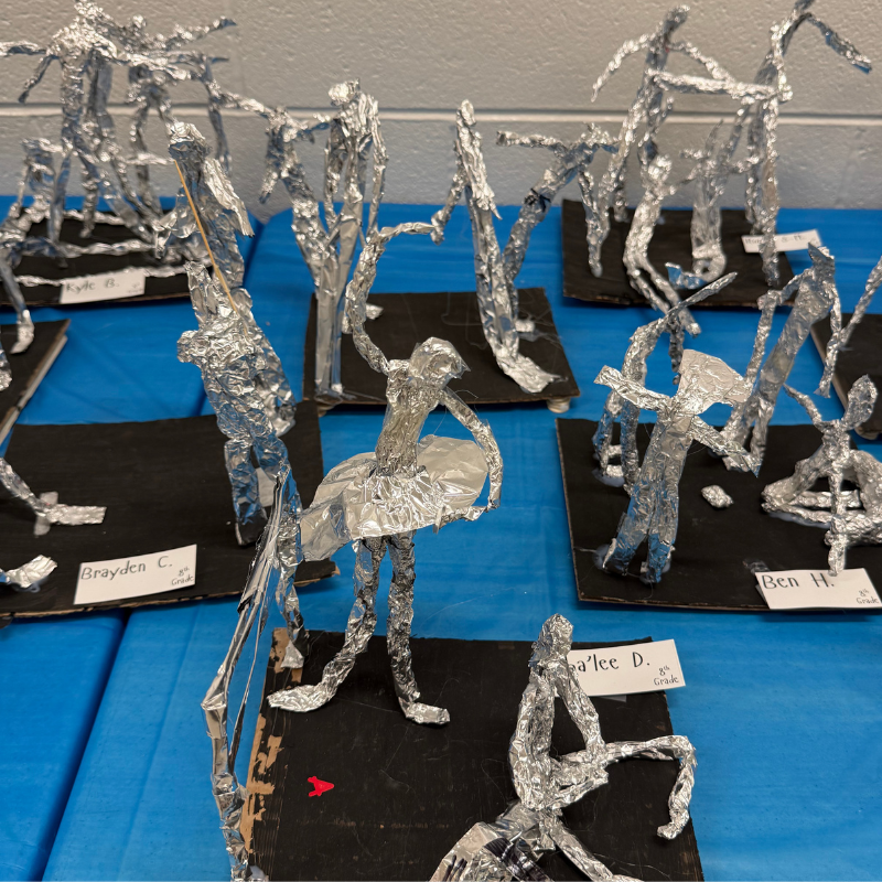A collection of aluminum foil sculptures depicting human figures in various poses, displayed on a blue tablecloth.