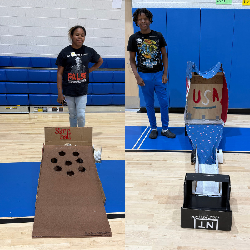 Two children stand next to homemade carnival games in a gymnasium.