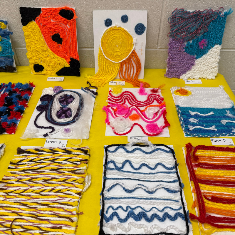 A display of colorful yarn art projects, including abstract designs and a seascape.