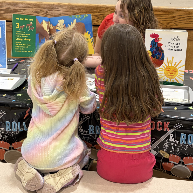 Two young girls and an adult look at children's books on a table decorated with musical notes and drums.