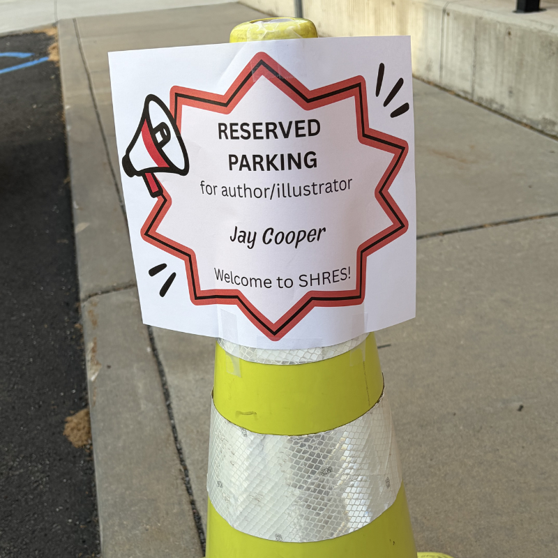 A sign on a yellow cone reads 'RESERVED PARKING for author/illustrator Jay Cooper Welcome to SHRES!'.