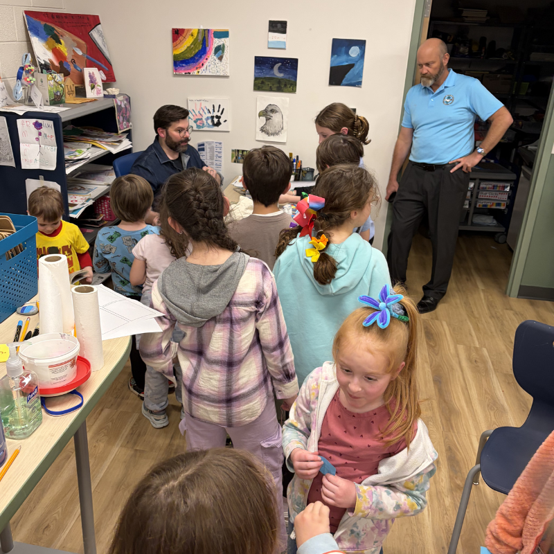 A group of children gather around a table with a man, while another man stands nearby in a blue shirt.