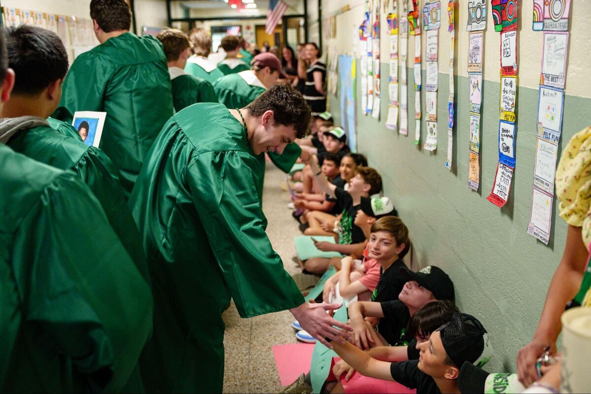 Graduates in green gowns interact in a school hallway decorated with student artwork.