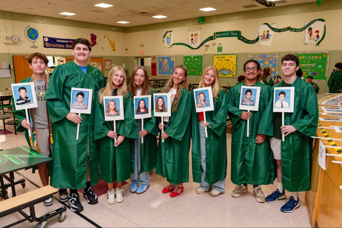 A group of students in green graduation gowns hold up individual portrait photos.