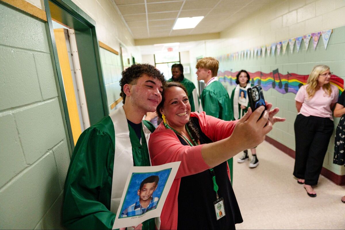 A woman takes a selfie with a smiling graduate in a green cap and gown in a school hallway.