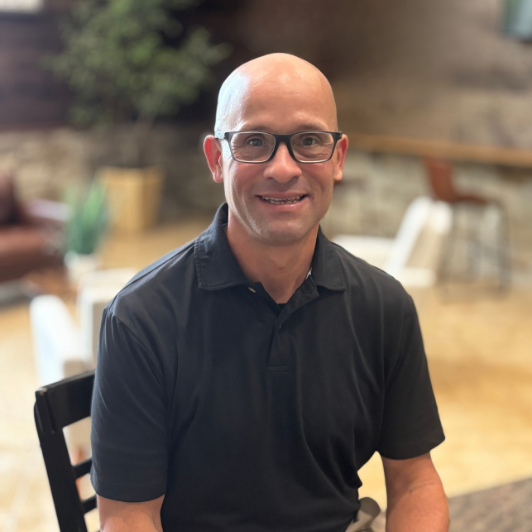 A smiling Johnny Lemen with glasses wearing a black polo shirt and sitting in a chair.