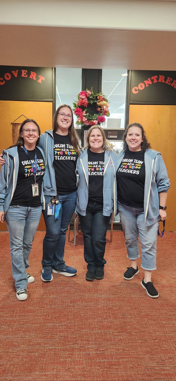 Four smiling female teachers stand together in a school hallway, wearing matching black t-shirts and blue zip-up hoodies.