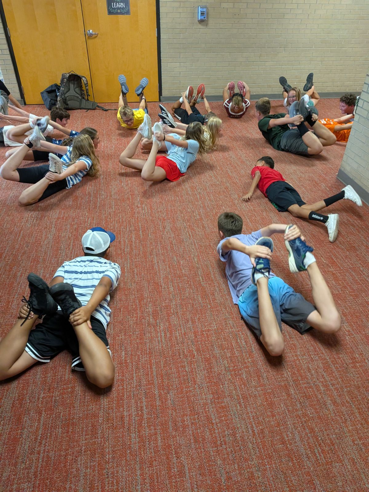 A group of cross country students lies on a red carpeted floor, stretching their legs in a school hallway.