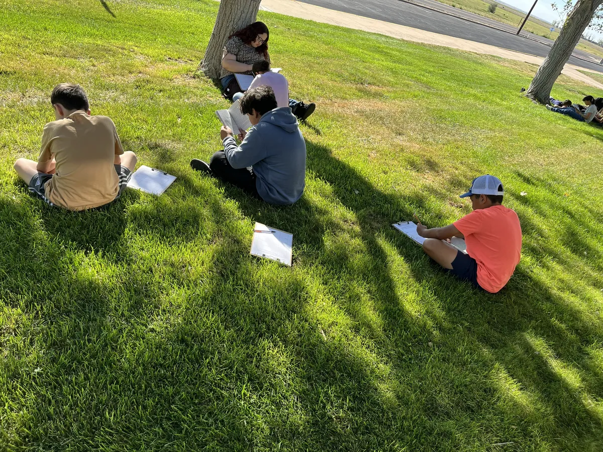 Students sit on the grassy lawn under tree shade, reading and writing outdoors on a sunny day.