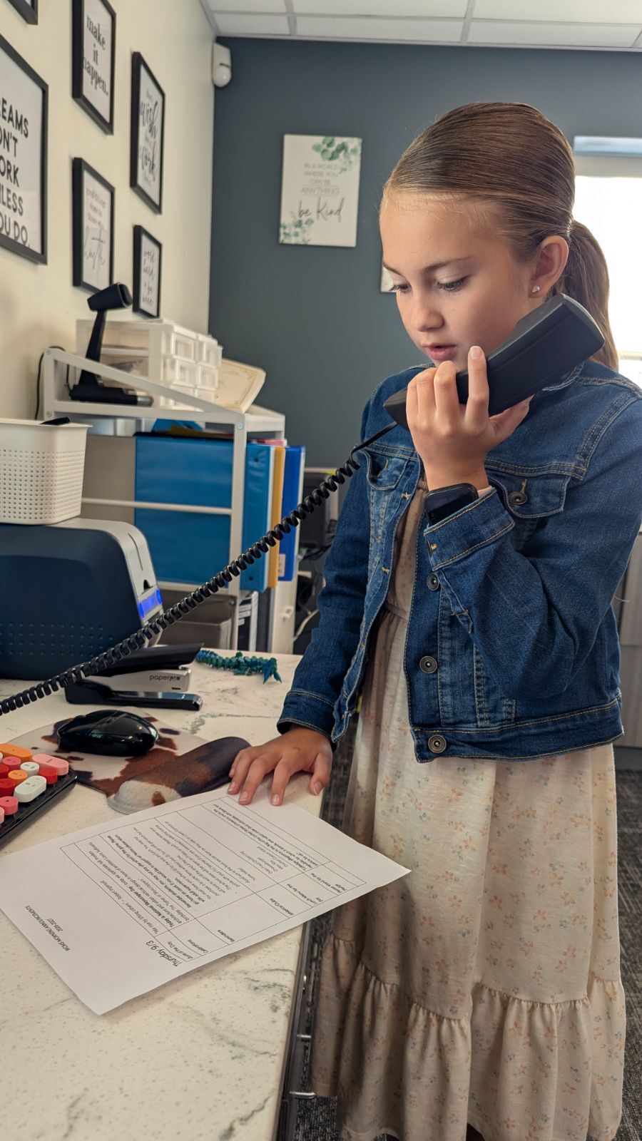 A young girl in a denim jacket and floral dress stands at a desk, talking on a black telephone while looking at a paper.