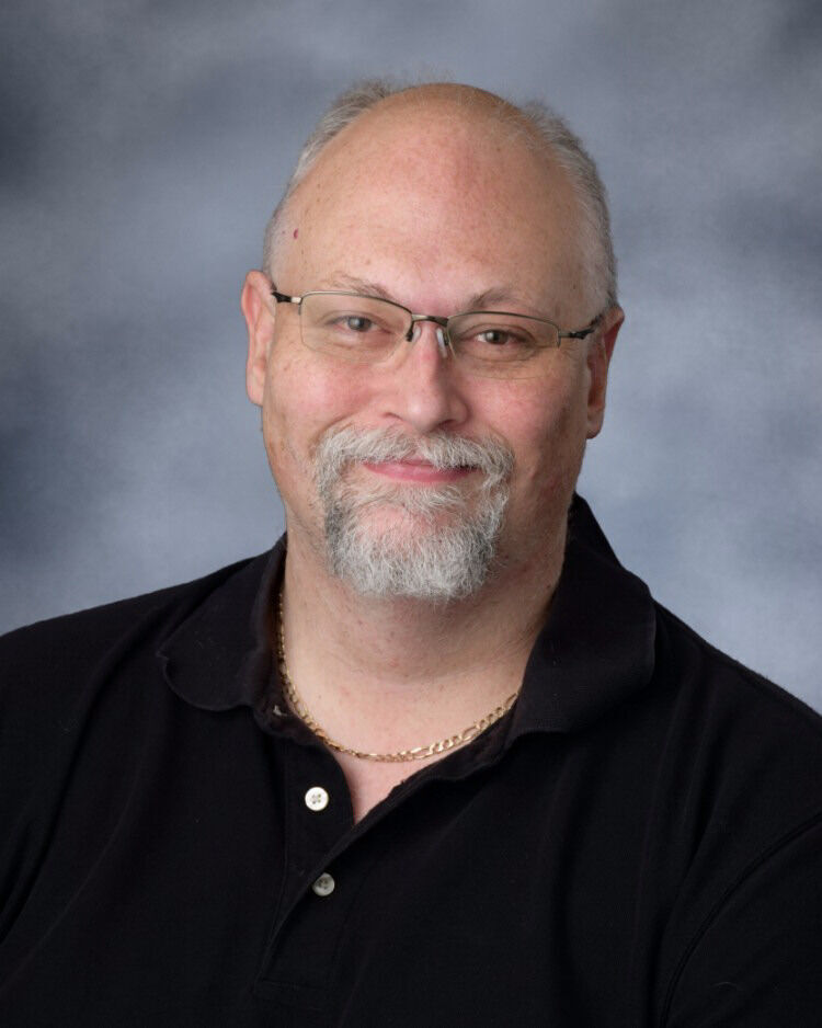 A middle-aged man with glasses, a graying beard, and a black polo shirt smiles against a gray backdrop.