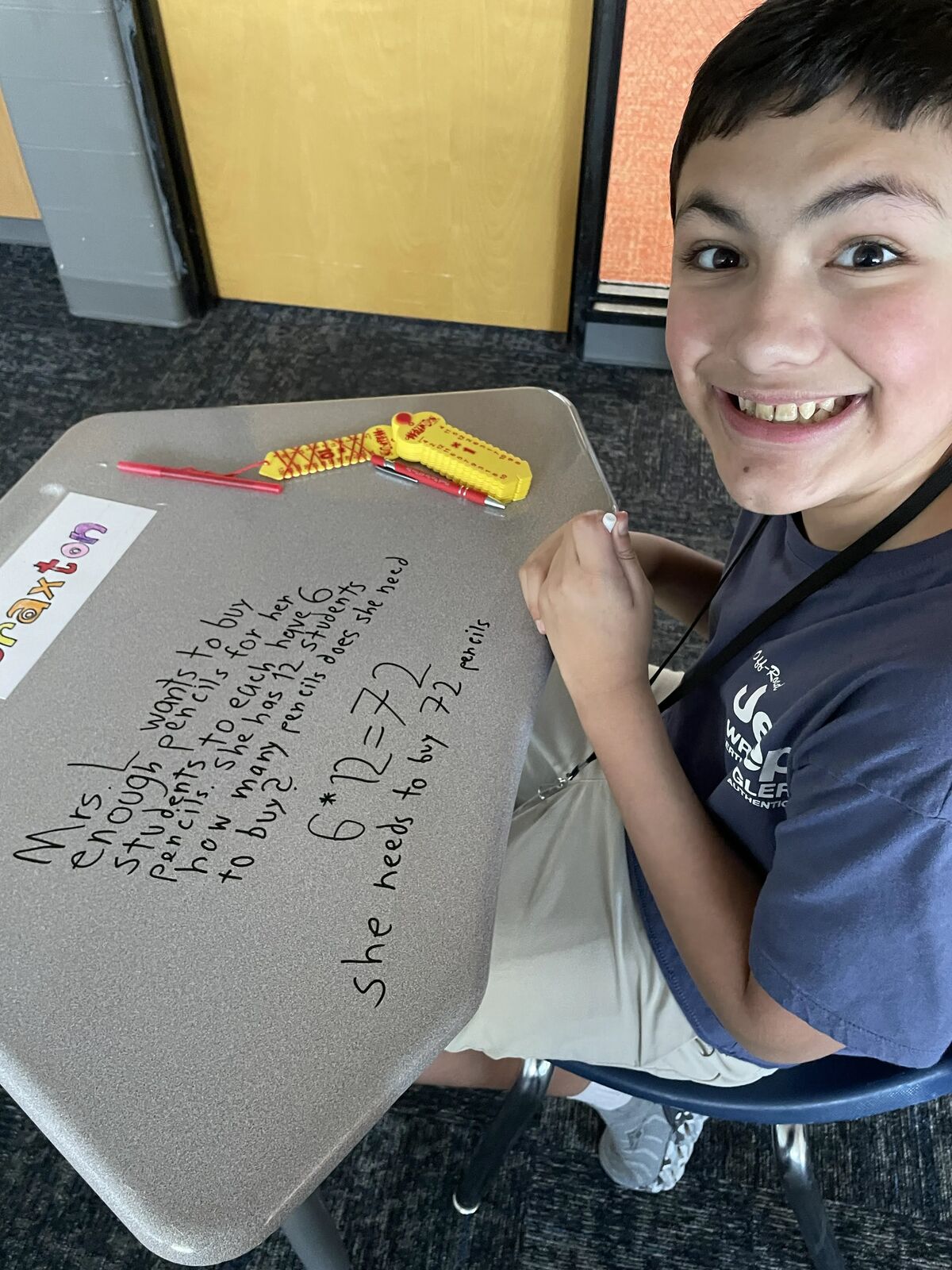 A smiling young boy sits at a school desk, proudly showing math equations written directly on the gray surface.