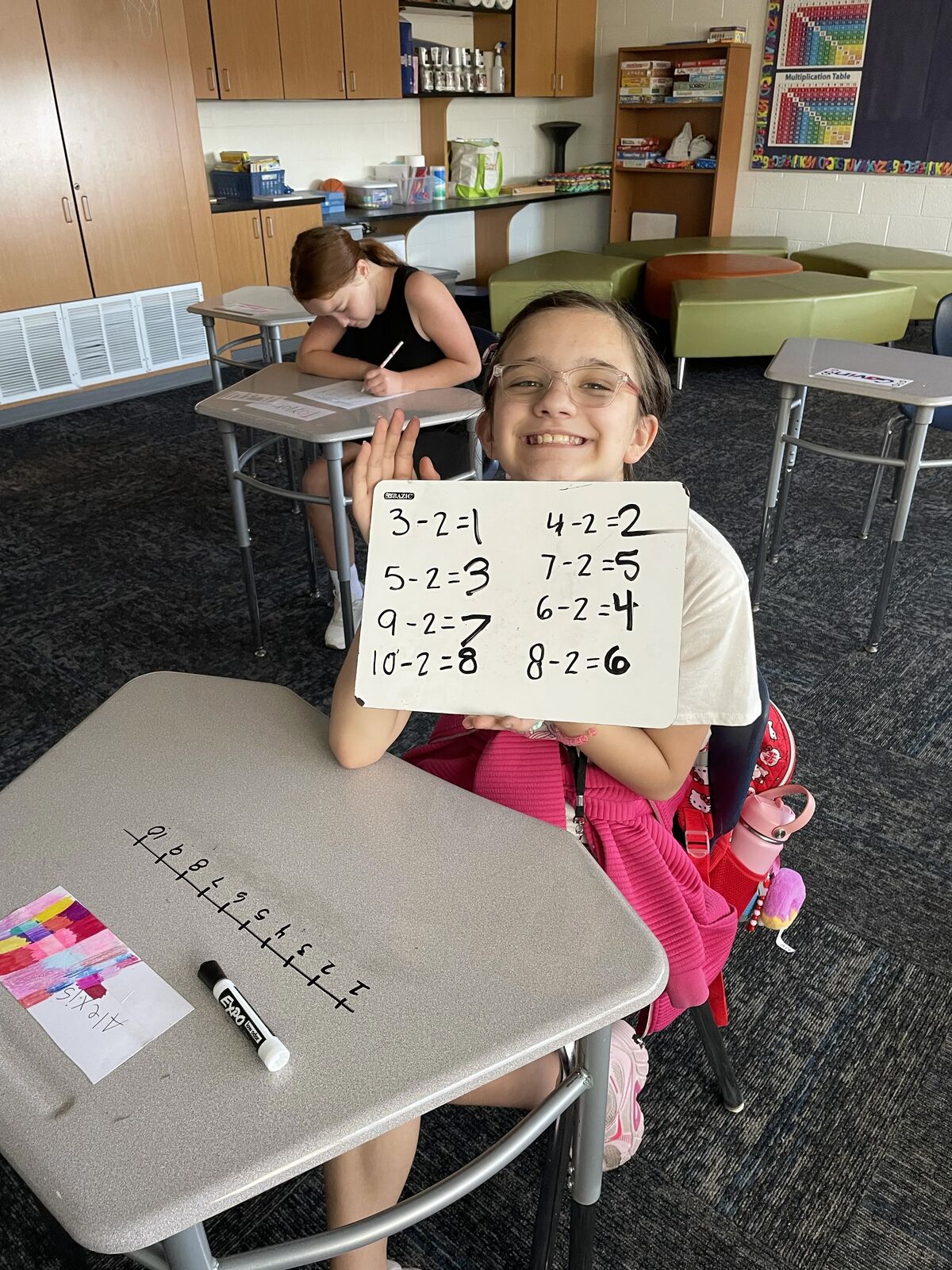 A smiling student holds up a whiteboard displaying math subtraction problems in a classroom.