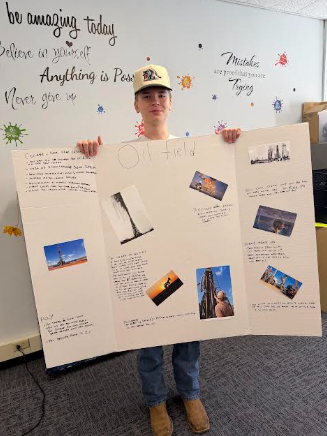 A young person stands in a room, holding a tri-fold poster board about an oil field, with inspirational quotes on the wall behind them.
