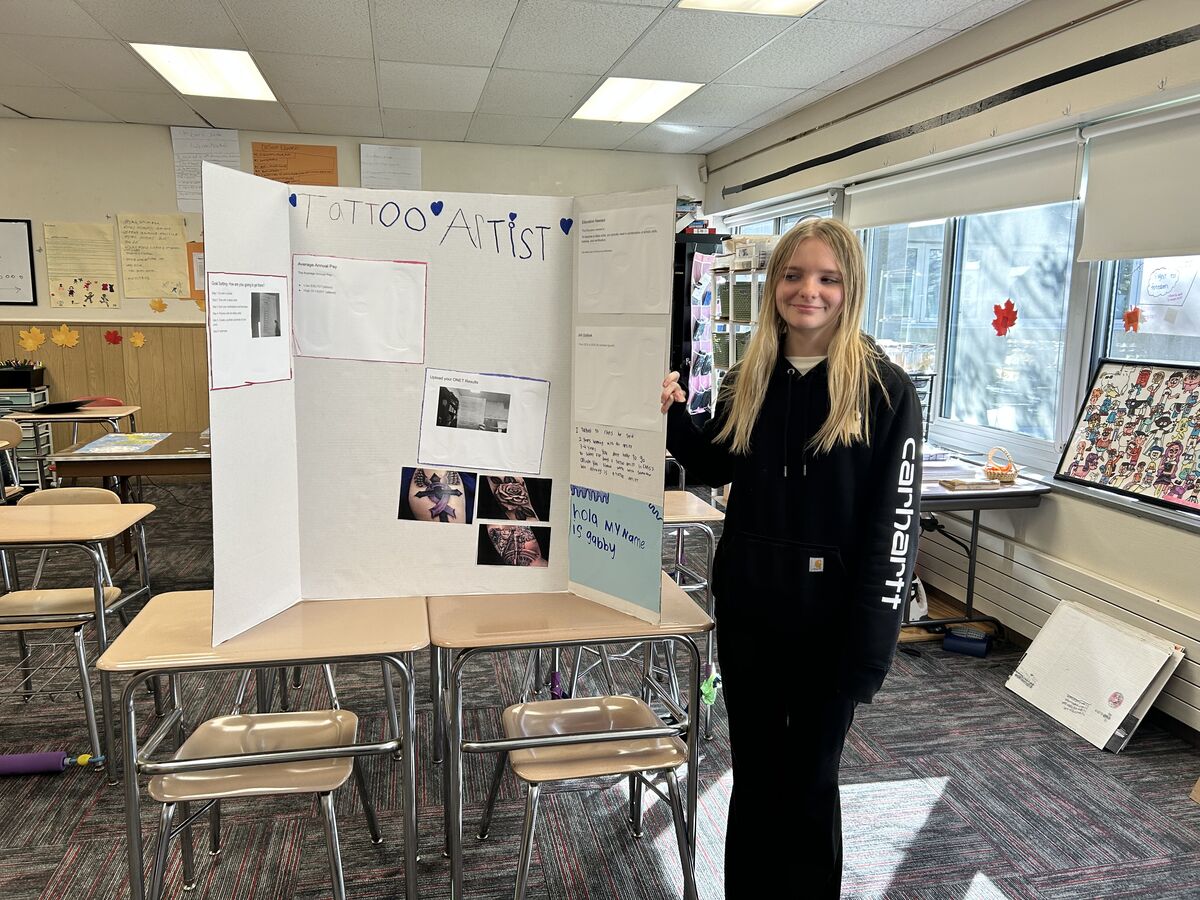 A student stands proudly next to a tri-fold display board titled "Tattoo Artist" inside a bright classroom.