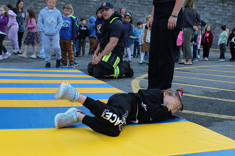 Young girl in sweats and sneakers covering her face with her hands, rolling on a mat with children and a firefighter looking on.