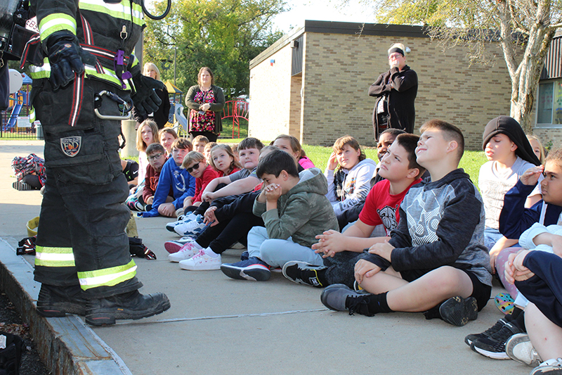 students seated on sidewalk outdoors, firefighters legs in foreground