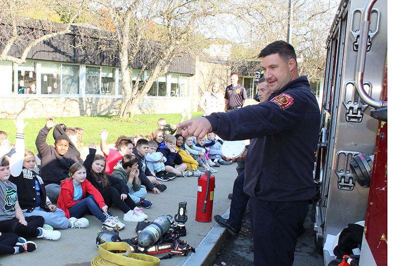 Children seated on sidewalk; firefighter pointing to someone, firefighting gear on sidewalk, edge of firetruck
