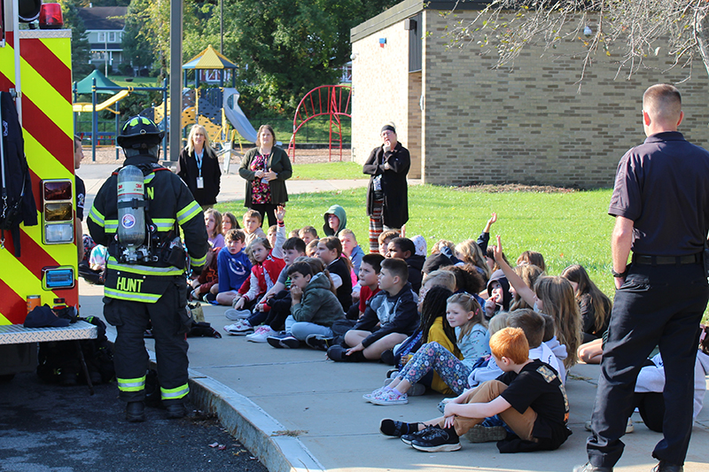 group of students seated on sidewalk, firetruck, firefighter in gear, other adults standing, grass, brick building