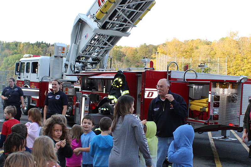 Small children standing in foreground, firefighters in uniform and ladder truck with ladder in air in background
