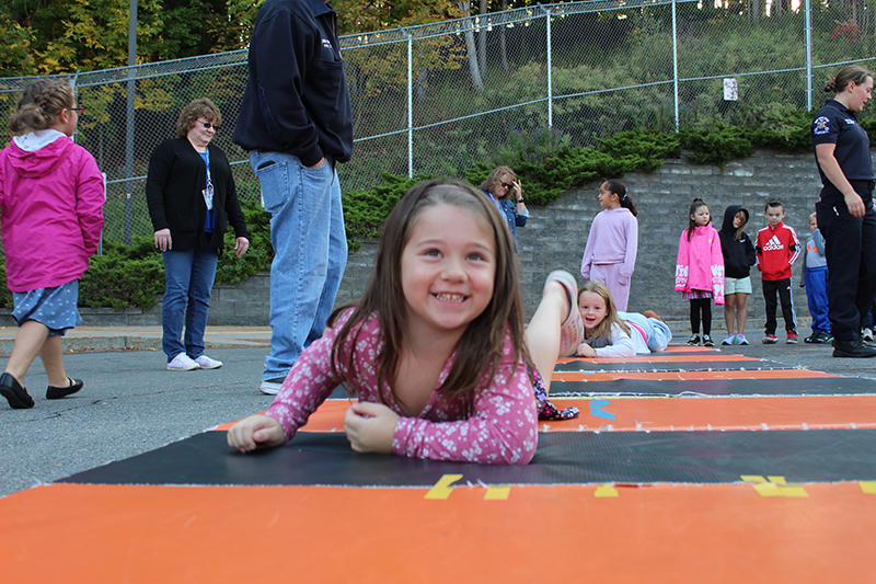 young girl look at camera, crawling on mat, other students in background, fence and shrubs in far back