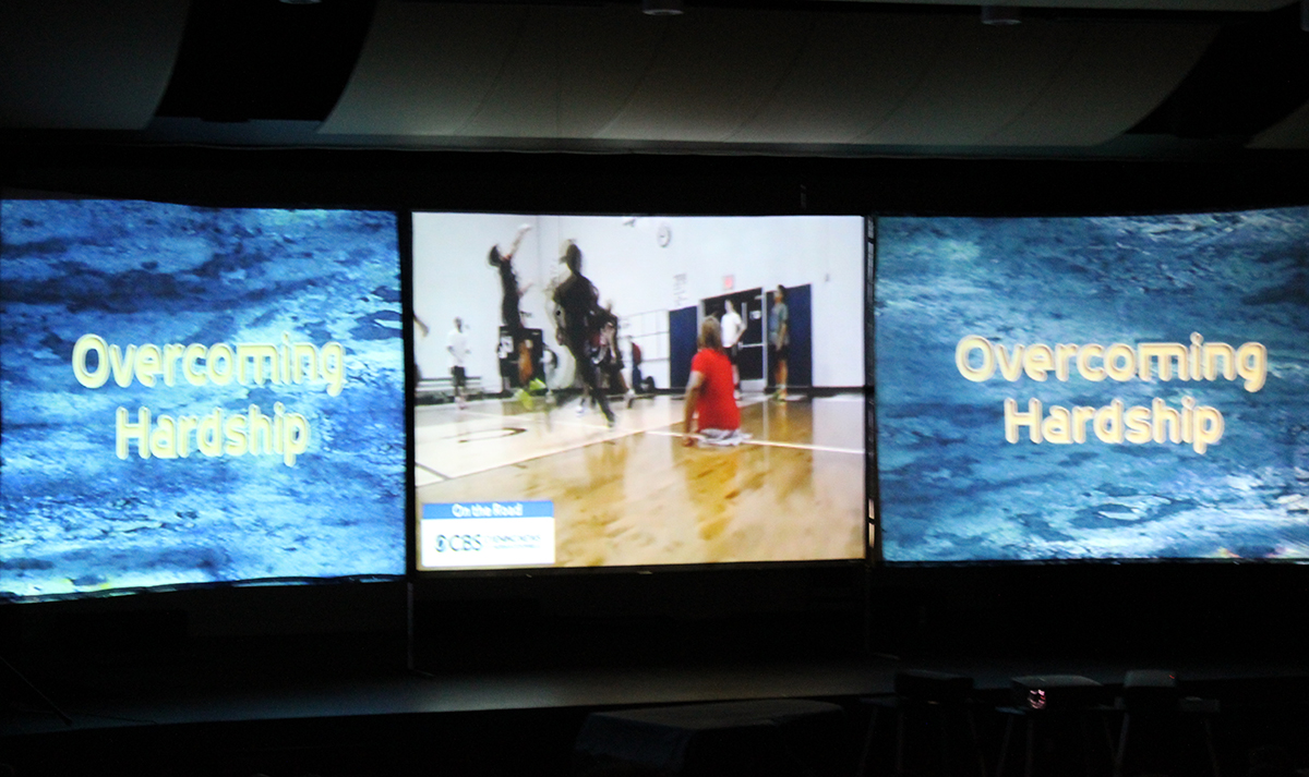 three screens on stage, outer two with words overcoming hardship, center screen with boys playing basketball