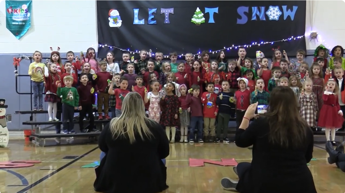 large number of young children standing on risers with big sign saying let it snow in the background