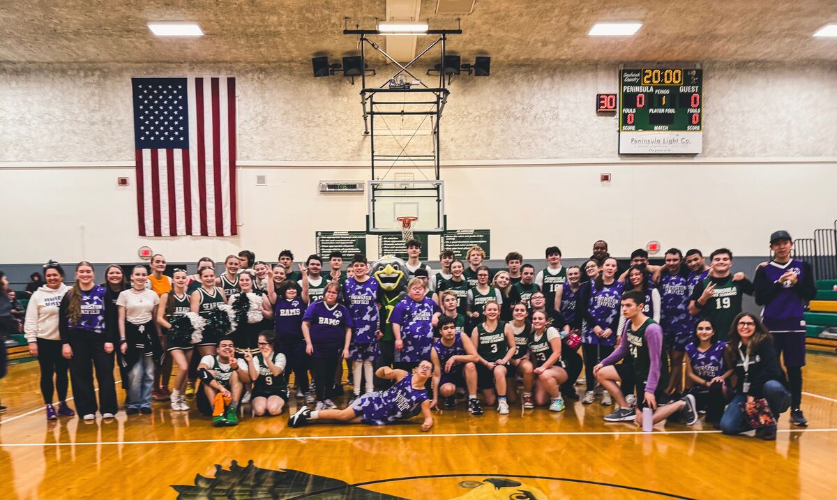 A heartwarming group photo taken in a high school gymnasium showcasing the unified spirit of Peninsula High School and North Thurston's basketball teams. Players from both teams, wearing purple and green jerseys respectively, are gathered together on the basketball court for a post-game photo. The school mascot, wearing a purple uniform, stands among the players, and the American flag hangs prominently on the wall behind them. The scoreboard and basketball hoop are visible in the background, while the polished wooden floor reflects the gymnasium lights. Students are displaying various poses of joy and camaraderie, including some playfully sitting or lying on the court.